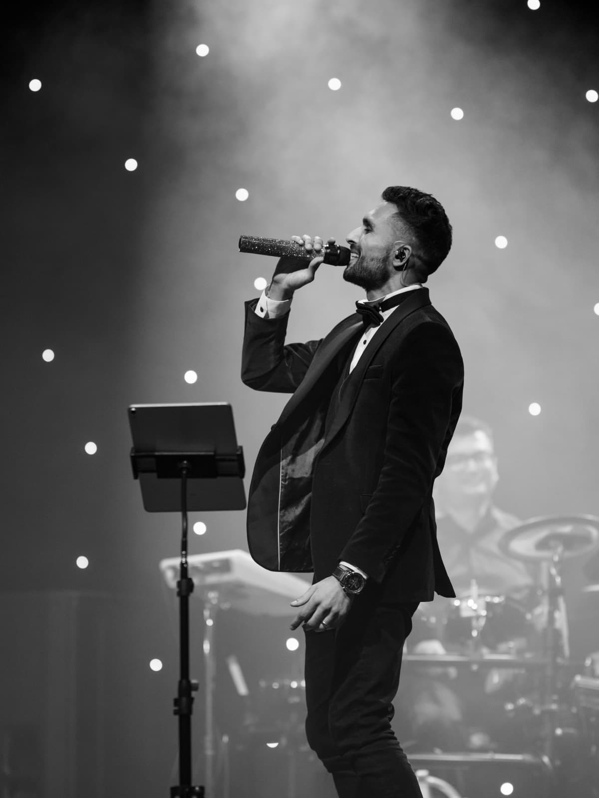 Paras singing into a microphone on stage in a velvet tuxedo, drummer behind him